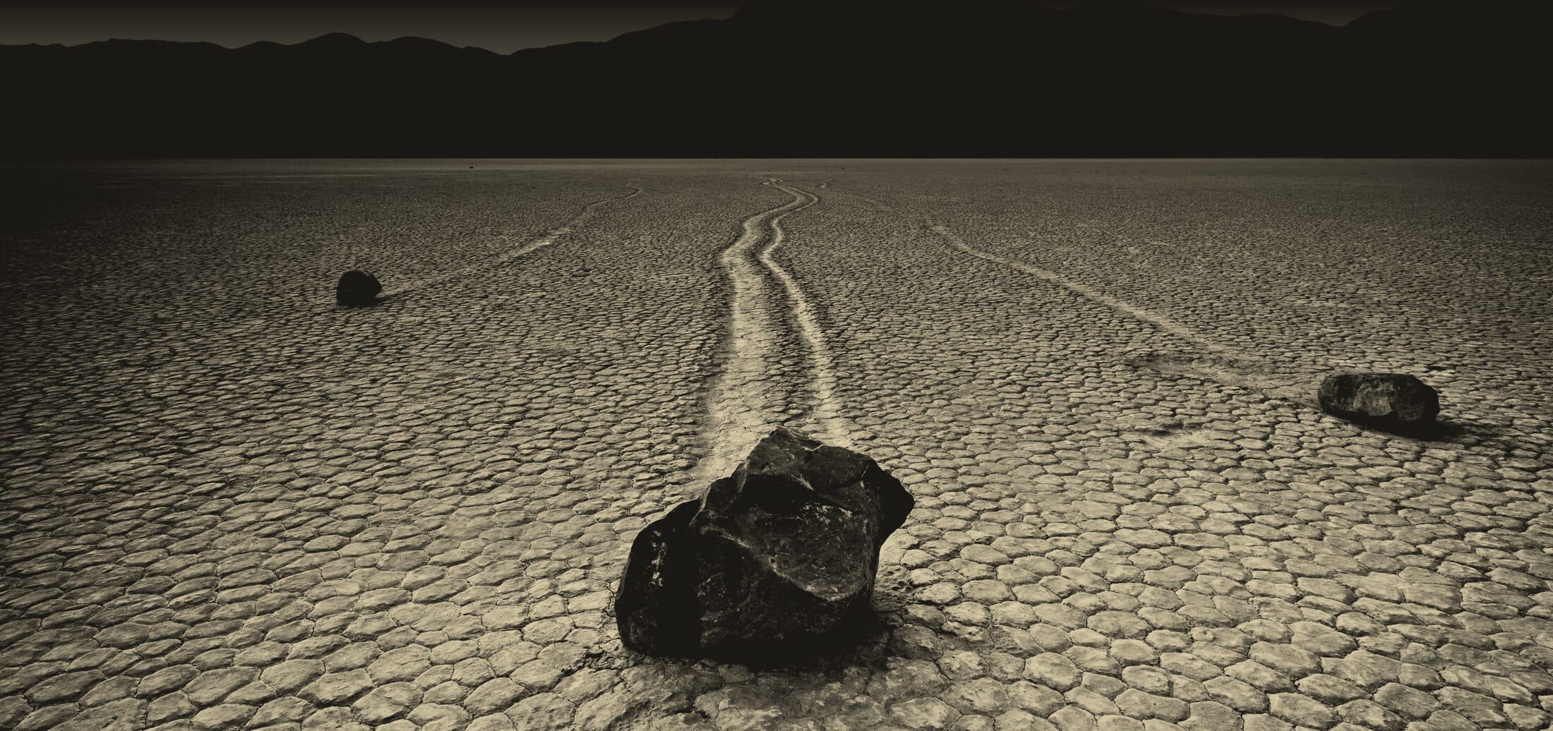 Une pierre sombre au crépuscule sur Racetrack Playa, dans la Vallée de la Mort, sa longue trace incurvée gravée dans la boue craquelée vers les montagnes en silhouette sur la crête.